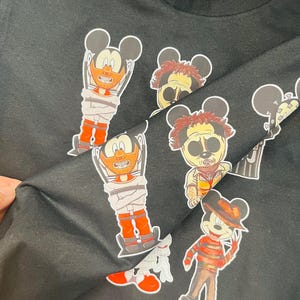 May include: Black crew neck t-shirt featuring cartoon character mashups. The graphics include Mickey Mouse characters dressed as horror movie villains, such as Freddy Krueger and Leatherface. The characters are in various poses and outfits.