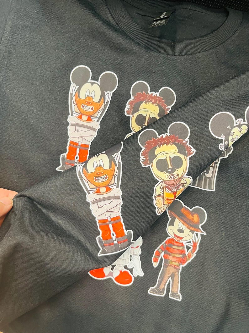 May include: Black crew neck t-shirt featuring cartoon character mashups. The graphics include Mickey Mouse characters dressed as horror movie villains, such as Freddy Krueger and Leatherface. The characters are in various poses and outfits.