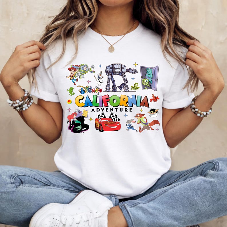 May include: White t-shirt featuring a colorful graphic of Disney characters and the words "California Adventure". The design includes images of characters from various Disney movies, such as Toy Story, Cars, and Monsters, Inc.