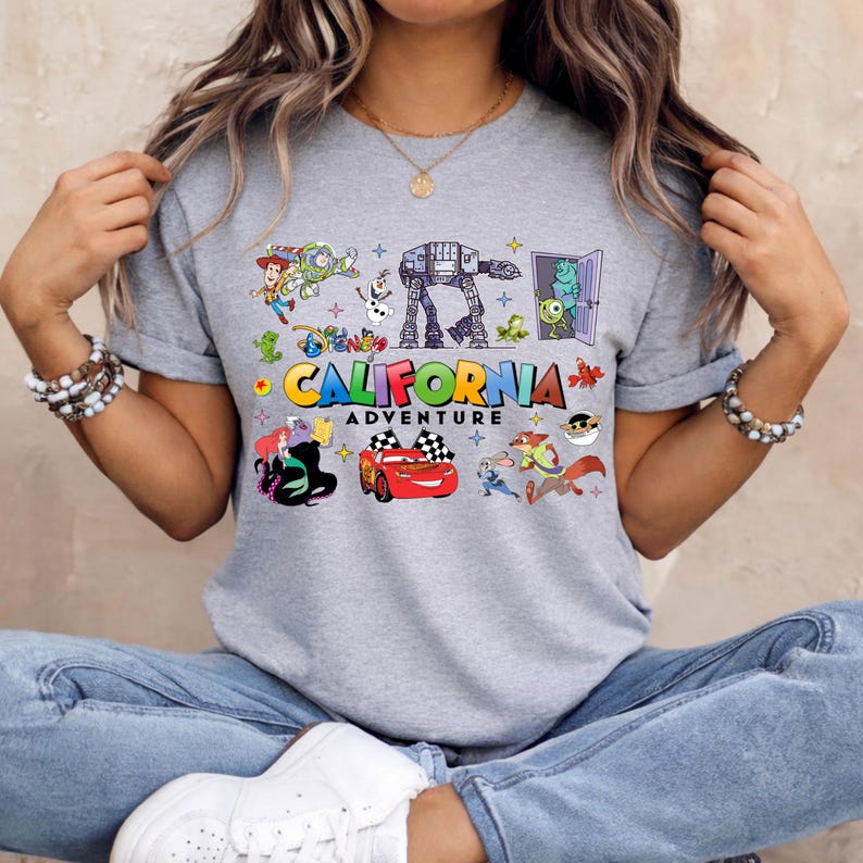 May include: A gray t-shirt featuring a colorful graphic design with the text "California Adventure". The design includes various cartoon characters and elements, such as a race car, an AT-AT walker, and other Disney-themed illustrations.