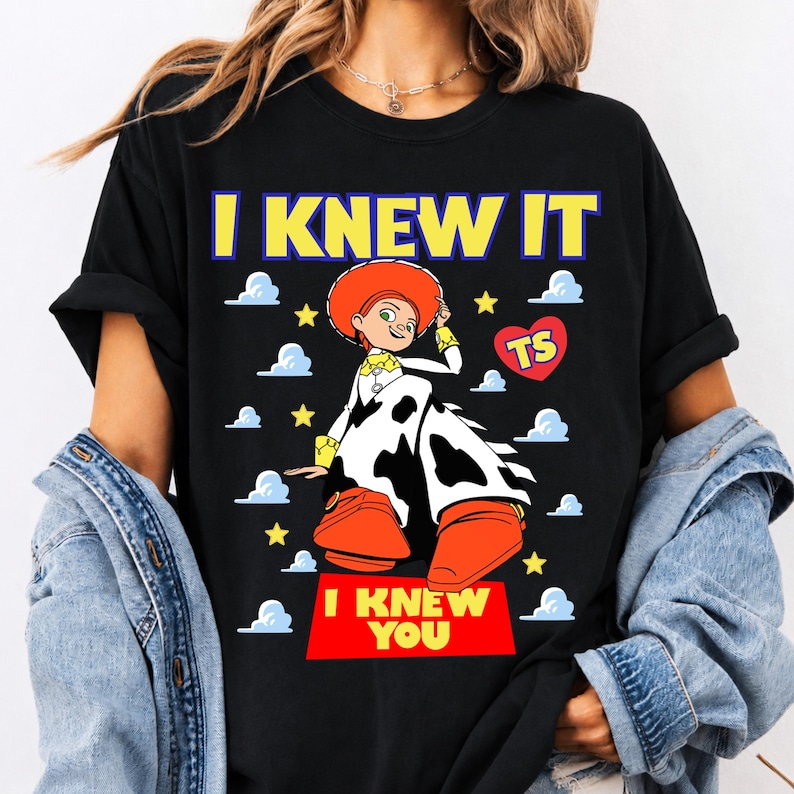 May include: A black t-shirt featuring a graphic of the character Jessie from Toy Story. The design includes the text I KNEW IT at the top and I KNEW YOU at the bottom, surrounded by yellow stars, blue clouds, and a red heart with the letters TS.
