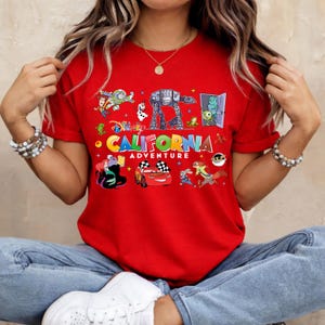May include: Red t-shirt with a colorful graphic featuring various Disney characters and the words "CALIFORNIA ADVENTURE". The design includes images of a race car, an AT-AT walker, and other animated figures.