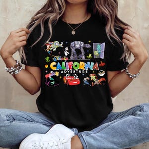 May include: Black t-shirt featuring colorful Disney characters and the text "California Adventure". The design includes characters from Toy Story, Monsters, Inc., Cars, and more. A fun and vibrant graphic tee.
