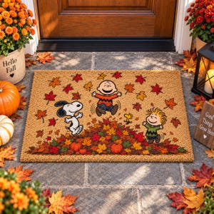 May include: A rectangular tan coir doormat featuring a colorful autumn scene with Snoopy, Charlie Brown, and Linus jumping into a pile of fallen leaves. The design includes orange, red, and yellow maple leaves, two small pumpkins, and Woodstock.