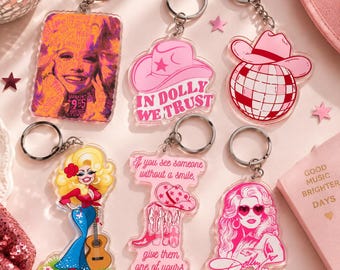 May include: Six acrylic keychains featuring Dolly Parton themes. Designs include a portrait with text, a pink cowboy hat with 'IN DOLLY WE TRUST', a disco ball with a hat, a cartoon figure with a guitar, boots with text, and a pink portrait with heart glasses.