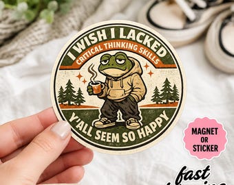 Pode incluir: Um autocolante ou íman circular com um sapo de desenho animado a usar um capuz bege e calças escuras, segurando uma caneca a fumegar. O texto diz I WISH I LACKED CRITICAL THINKING SKILLS Y'ALL SEEM SO HAPPY. O design inclui pinheiros e estrelas.