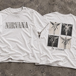 Puede incluir: Una camiseta blanca de manga corta con la palabra NIRVANA impresa en negro en la parte delantera. La parte trasera muestra un gráfico de cuatro paneles cuadrados con figuras aladas y el texto IN UTERO impreso en negro bajo los paneles.