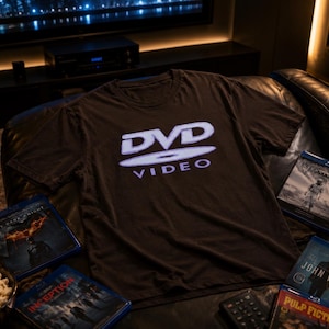 Pode incluir: Uma t-shirt preta de manga curta com o logótipo branco DVD Video impresso no peito. A peça é exibida ao lado de várias caixas de filmes em Blu-ray, incluindo The Dark Knight, Interstellar, John Wick, Inception e Pulp Fiction.