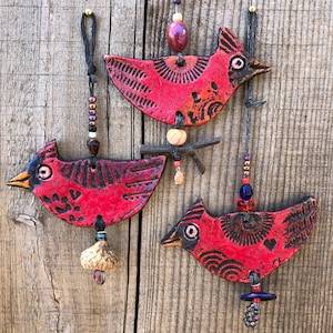 May include: Three red cardinal bird ornaments with intricate details and black accents. Each ornament is suspended from a string with beads and charms.