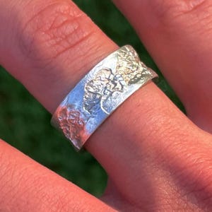 Könnte beinhalten: Ein silberner Ring mit strukturierter Oberfläche und floralem Design. Der Ring wird an einem Finger getragen und zeigt seine komplizierten Details und seine Handwerkskunst. Das Design des Rings weist eine Mischung aus glatten und rauen Texturen auf.
