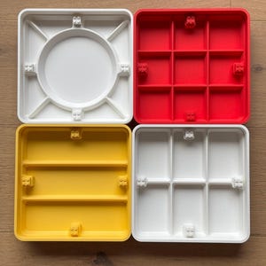 May include: Four colourful plastic trays in white, red, yellow, and white. Each tray has a unique design, including a circular centre, divided compartments, and raised edges. The trays are designed to interlock with each other.