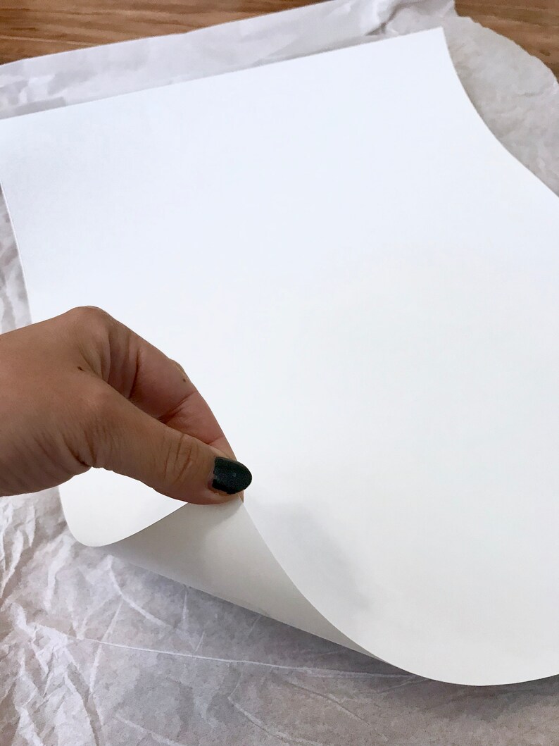 May include: A hand lifting a corner of a blank, white sheet of paper, revealing the underside. The paper is smooth and bright, with a slight curve where it is being lifted. The background is a textured, off-white surface.