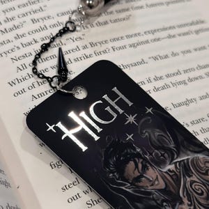 May include: A black bookmark with a silver chain and a black tassel. The bookmark features a silver metallic design that says "HIGH" with stars. The bookmark also features a black and white illustration of a man with a tattoo.