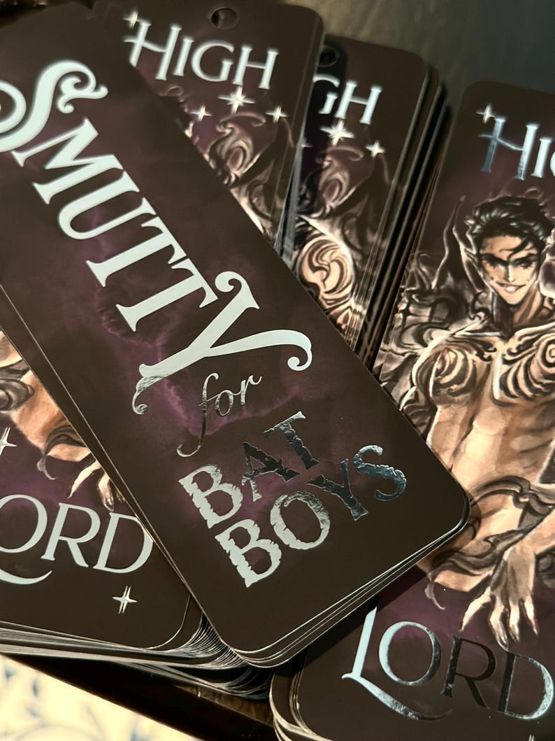 May include: A stack of dark brown bookmarks with silver foil lettering. The text on the bookmark reads "Smutty for Bad Boys" and "High Lord".