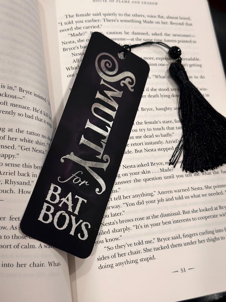 May include: A black bookmark with silver lettering that reads "Smutty for Bat Boys". The bookmark has a black tassel.