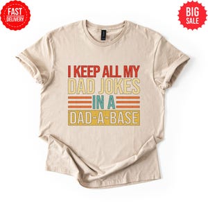 Op de afbeelding: Beige t-shirt met de tekst "I KEEP ALL MY DAD JOKES IN A DAD-A-BASE" in rood, geel en blauw. Het shirt heeft een vintage, retro design. Inclusief labels "FAST DELIVERY" en "BIG SALE".