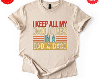 May include: Beige t-shirt with the text "I KEEP ALL MY DAD JOKES IN A DAD-A-BASE" in red, yellow, and blue. The shirt has a vintage, retro design. Includes "FAST DELIVERY" and "BIG SALE" labels.