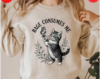 Op de afbeelding: Beige sweatshirt met een zwart-witte illustratie van een kat die een vlinder vasthoudt. De tekst "RAGE CONSUMES ME" staat boven de kat. Bloemenelementen omringen de kat en de tekst. De sweatshirt heeft lange mouwen.