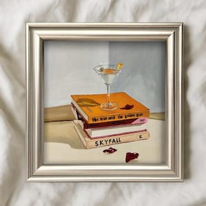 May include: A framed painting featuring a martini glass with an olive and lemon twist, atop a stack of books. The books are titled "The Man with the Golden Gun" and "Skyfall". A red object is on the table.