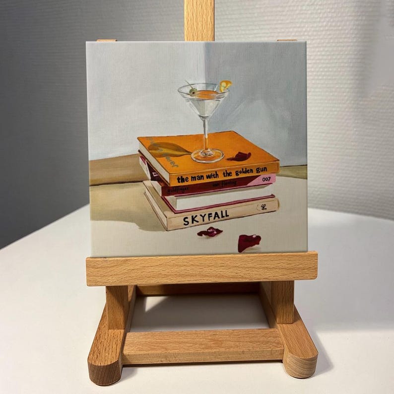 May include: A still-life painting on a small easel depicts a martini glass atop a stack of books. The books are titled "the man with the golden gun" and "SKYFALL". The martini has an olive and lemon peel garnish. The books are orange, pink, and white.