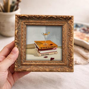 May include: A framed painting featuring a martini glass with a lemon garnish atop a stack of books. The books are titled "the man with the golden gun" and "SKYFALL". The frame is ornate with a gold finish.