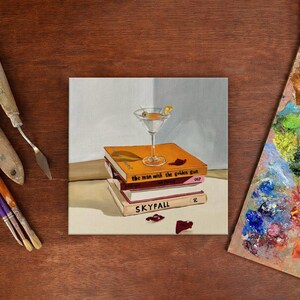 May include: An oil painting depicts a martini glass with a lemon twist atop a stack of books. The books are titled "the man with the golden gun" and "SKYFALL". The painting is on a white canvas, with a wooden background.
