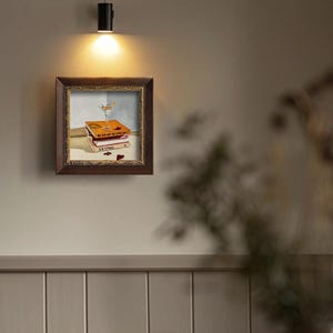 May include: A framed artwork depicts a still life with books and a cocktail glass. The books are stacked, with titles visible. A martini glass with a lemon garnish sits atop the books. The frame is dark brown.