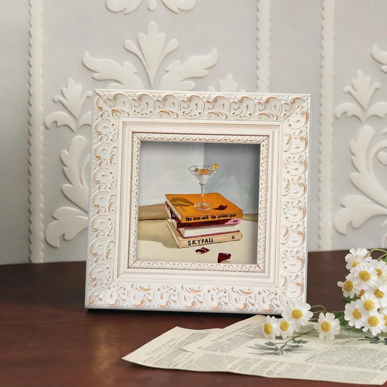 May include: A framed painting of a martini glass atop a stack of books titled "The Man with the Golden Gun" and "Skyfall." The frame is white with ornate detailing. A sprig of daisies and a newspaper are in the foreground.