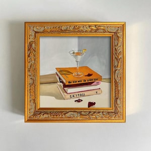 May include: A framed painting depicts a martini glass atop a stack of books. The books are titled "the man with the golden gun" and "SKYFALL". The painting has a gold ornate frame and a neutral background.