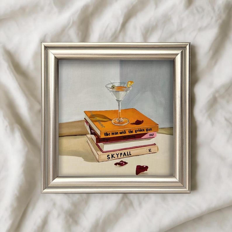May include: A framed painting featuring a martini glass with an olive and lemon twist, atop a stack of books. The books are titled "The Man with the Golden Gun" and "Skyfall". A red object is on the table.
