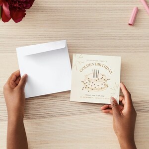 May include: A cream-colored birthday invitation with gold text and a cake illustration. The invitation is held with a white envelope on a light wood surface. A vase with a red flower and two pink crayons are also visible.