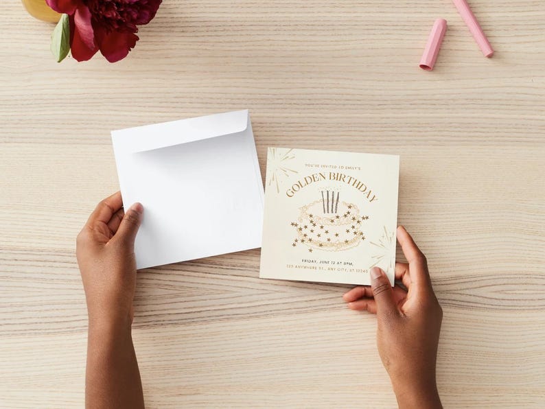 May include: A cream-colored birthday invitation with gold text and a cake illustration. The invitation is held with a white envelope on a light wood surface. A vase with a red flower and two pink crayons are also visible.