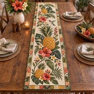 May include: A rectangular quilted table runner featuring a tropical pattern of yellow pineapples, red hibiscus flowers, white plumeria, green palm leaves, and a hummingbird, bordered by a patchwork design of small squares in earth tones.