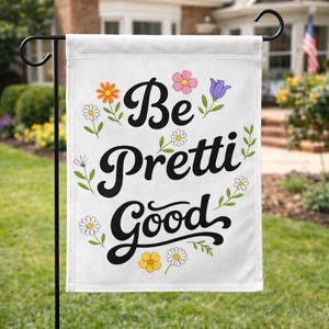 May include: White garden flag with the words "Be Pretti Good" in black script, surrounded by colorful flowers and green leaves. The flag is hanging on a black metal stand in a grassy yard.