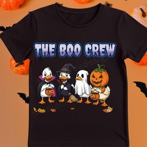 Op de afbeelding: Zwart T-shirt met de tekst "THE BOO CREW" in blauwe en witte letters. De afbeelding toont vier cartoon eenden verkleed in Halloween kostuums: een vampier, een heks, een spook en een pompoen. Elke eend houdt een snoepemmer vast.