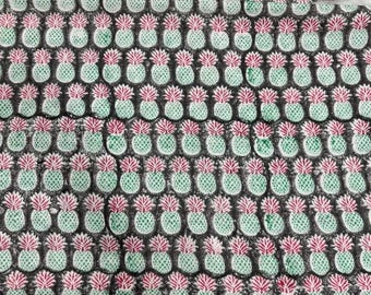 May include: A fabric with a repeating pattern of green and pink pineapples on a dark gray background. The pineapples are evenly spaced, creating a visually appealing and tropical design. The fabric appears to be cotton or a similar material.