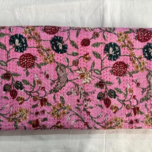 May include: A pink floral quilt with a repeating pattern of flowers and leaves in shades of red, green, yellow, and white. The quilt is hand-stitched with white thread, creating a textured surface. The overall theme is a floral home decor item.