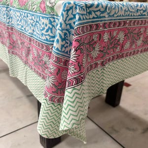 May include: A colorful floral patterned tablecloth. The design features a combination of pink, blue, and green hues with intricate floral and geometric patterns. The tablecloth is draped over a dark wooden table, showcasing its design.