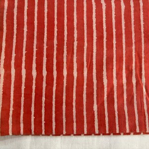 May include: Folded fabric with a repeating pattern of vertical white stripes on a red background. The stripes have a slightly uneven, hand-painted appearance, creating a textured look. The fabric is likely cotton or linen.
