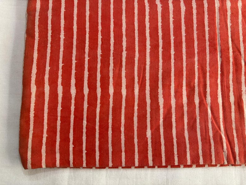 May include: Folded fabric with a repeating pattern of vertical white stripes on a red background. The stripes have a slightly uneven, hand-painted appearance, creating a textured look. The fabric is likely cotton or linen.