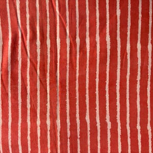 May include: Close-up of a fabric with a repeating pattern of vertical white stripes on a red background. The stripes have a slightly uneven, hand-drawn appearance, creating a textured look. The fabric appears to be slightly wrinkled.