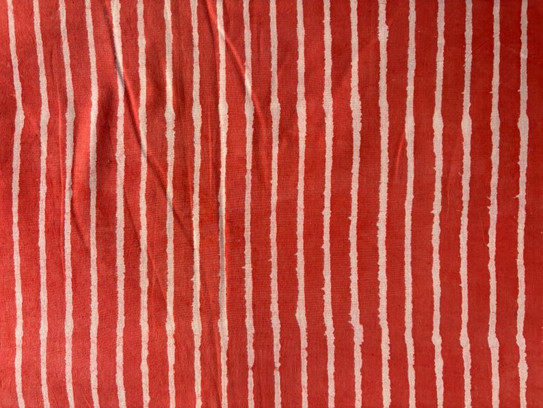 May include: Close-up of a fabric with a repeating pattern of vertical white stripes on a red background. The stripes have a slightly uneven, hand-drawn appearance, creating a textured look. The fabric appears to be slightly wrinkled.