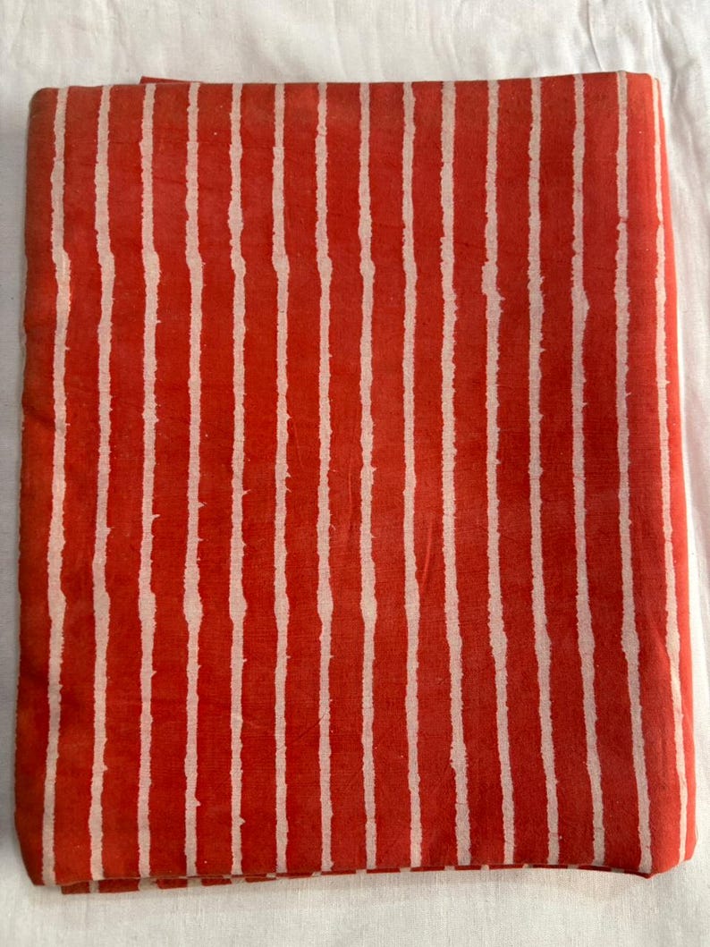 May include: Folded fabric with a vibrant red background and vertical white stripes. The stripes have a slightly irregular, hand-drawn appearance, creating a textured look. The fabric is likely cotton or a similar material.