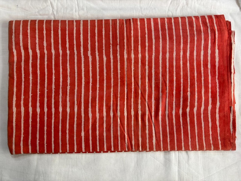 May include: Folded fabric with a repeating pattern of vertical white stripes on a red background. The fabric is folded and appears to be made of a soft material. The stripes are evenly spaced.