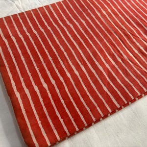 May include: Folded fabric with a repeating pattern of white stripes on a red background. The fabric appears to be cotton or a similar material, with a slightly textured surface. The stripes are uneven, adding to the handmade aesthetic.