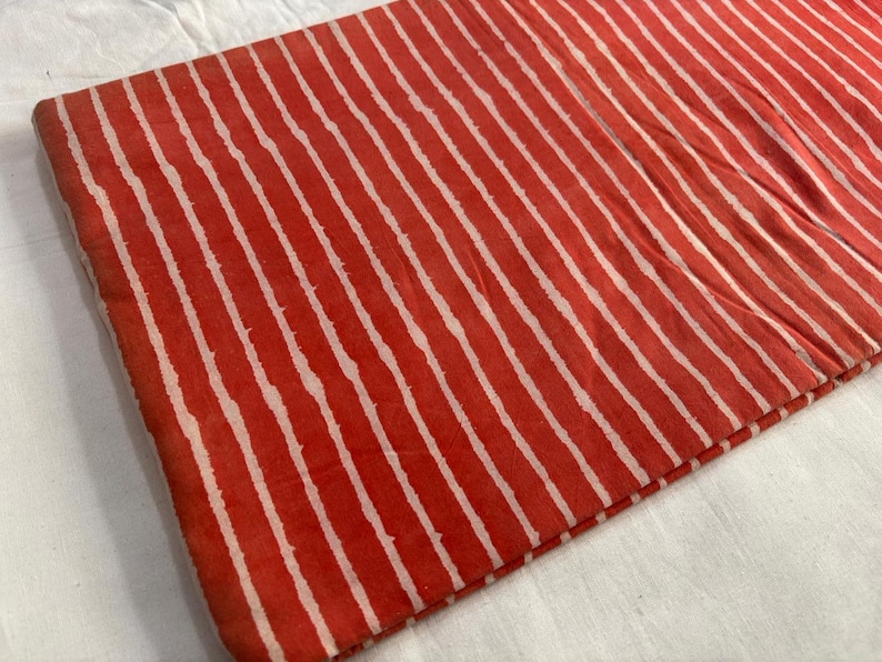 May include: Folded fabric with a repeating pattern of white stripes on a red background. The fabric appears to be cotton or a similar material, with a slightly textured surface. The stripes are uneven, adding to the handmade aesthetic.