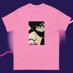 Puede incluir: Camiseta rosa con una ilustración de estilo pop art de un rostro femenino. El gráfico muestra una máscara negra, un ojo verde y labios rojos. La camiseta está hecha de un material suave y cómodo.