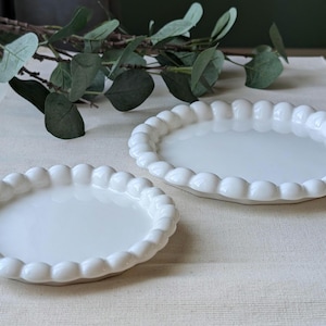 May include: Two white ceramic oval plates with a beaded edge. The plates are on a cream-coloured surface, with green leaves in the background. The plates are of different sizes.