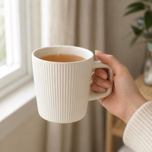 May include: A matte off-white ceramic mug featuring a vertical ribbed texture on the exterior. The mug has a sturdy handle and is filled with a warm, steaming amber-colored liquid, creating a simple and functional vessel for hot beverages.