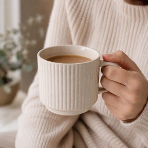 May include: A cream-coloured ceramic mug with a speckled finish and vertical ribbed texture. The mug features a sturdy side handle and is filled with a light brown liquid, likely coffee or tea, showing a smooth surface within the cylindrical vessel.